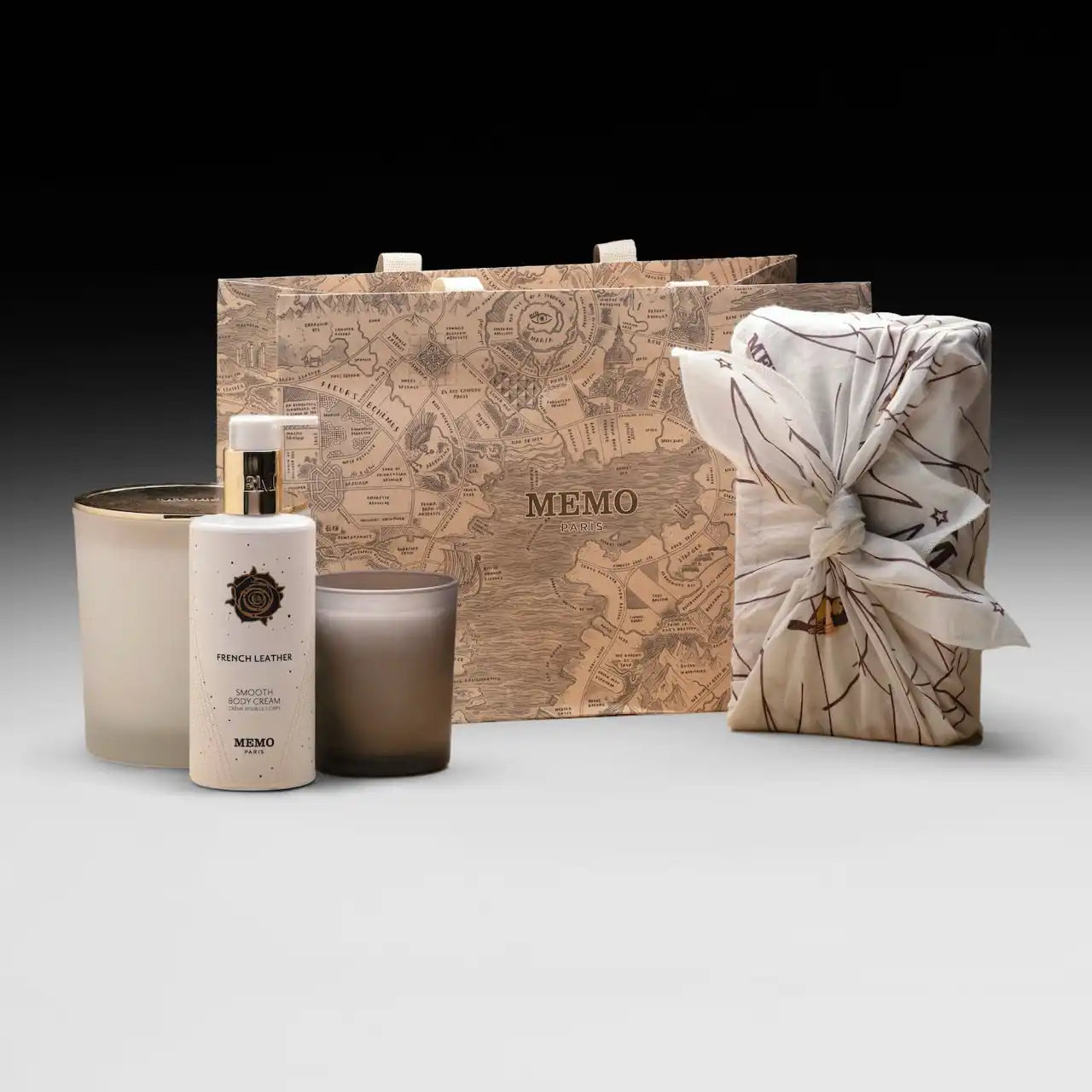A collection of Memo Paris brand products, including a shopping bag, a wrapped gift, a bottle of body lotion or fragrance, and two candles.