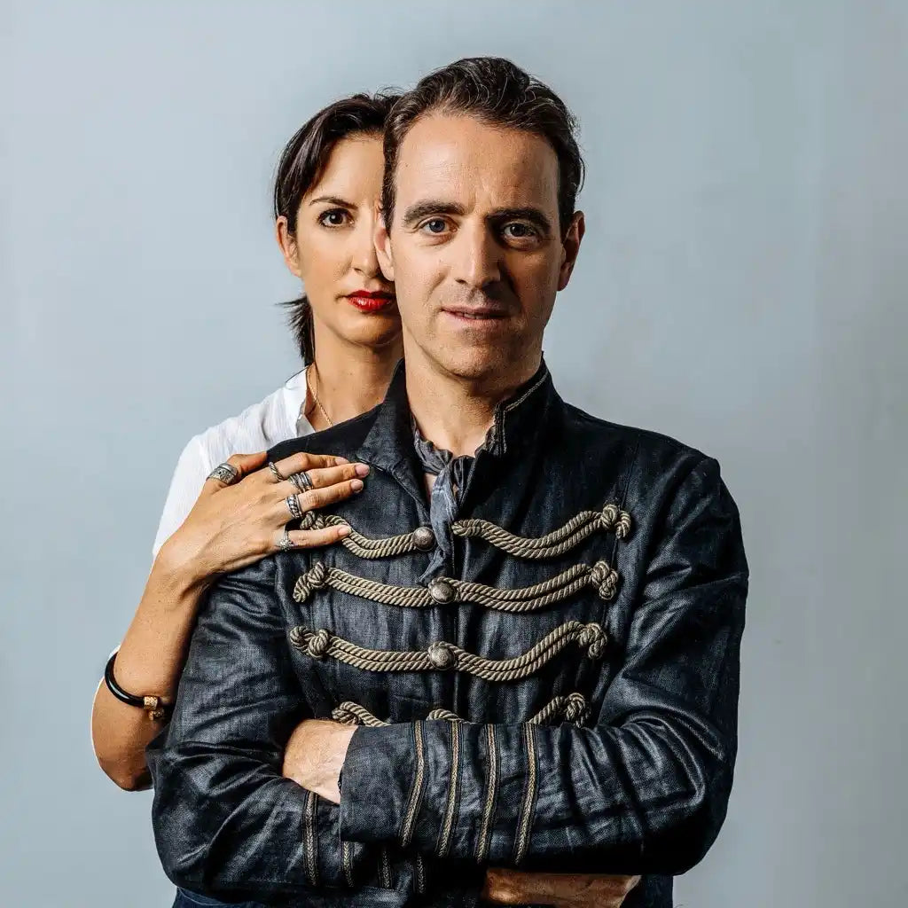 A man and a woman posing together, with the man in the foreground wearing an ornate dark jacket and the woman behind him with her hand on his shoulder.