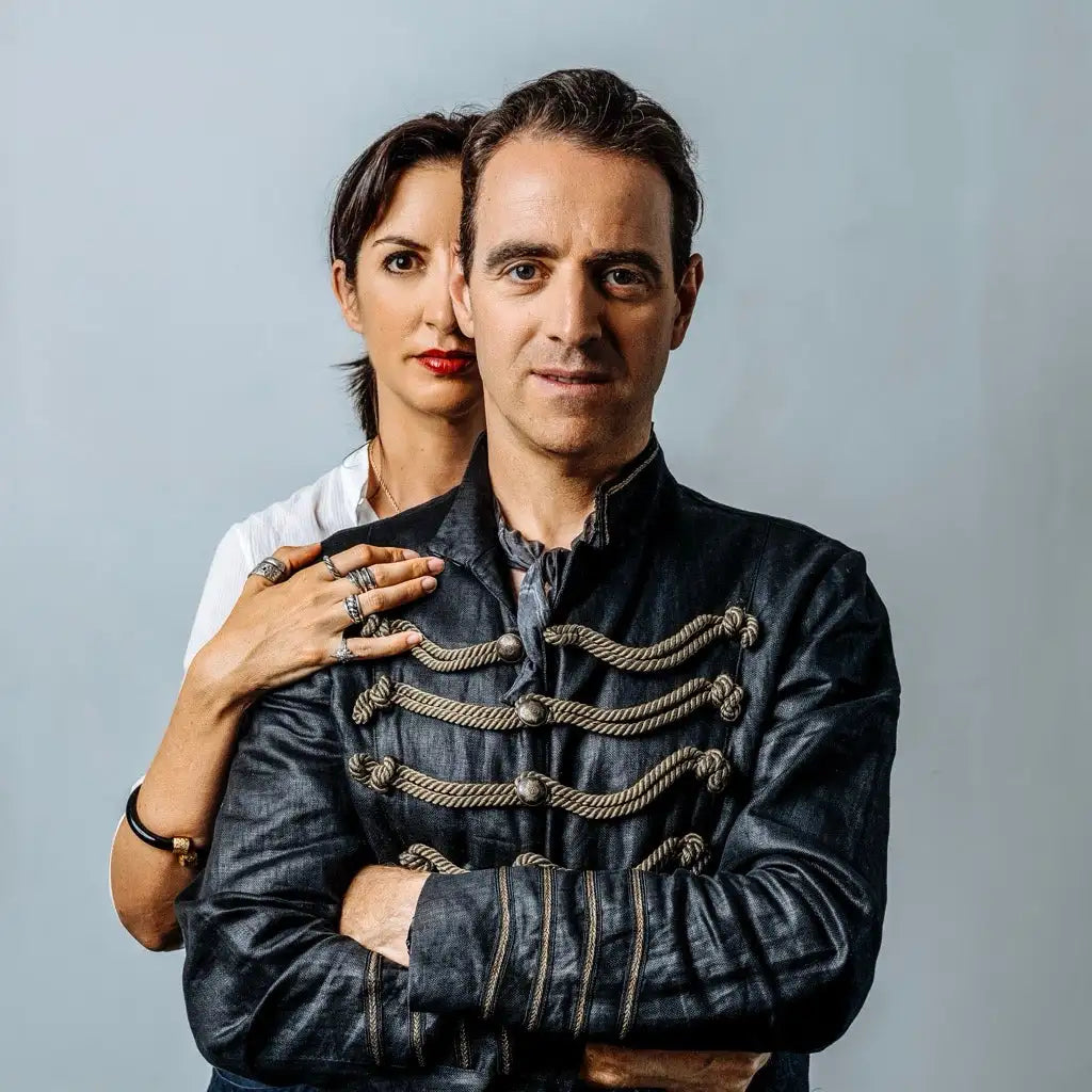A man and a woman posing together, with the man in the foreground wearing an ornate dark jacket and the woman behind him with her hand on his shoulder.