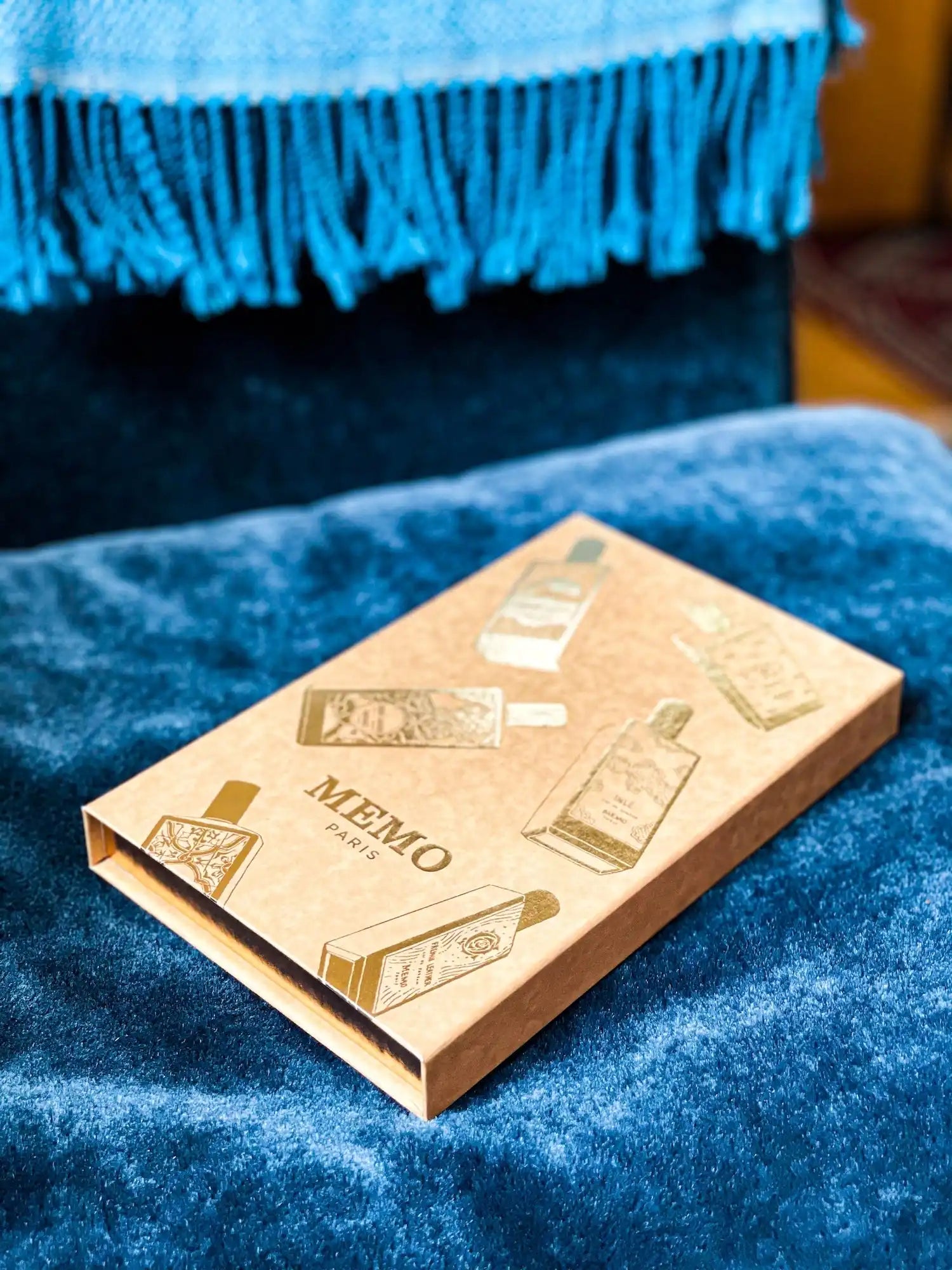 A rectangular, light brown perfume box with gold illustrations of perfume bottles and ’MEMO PARIS’ in the center.