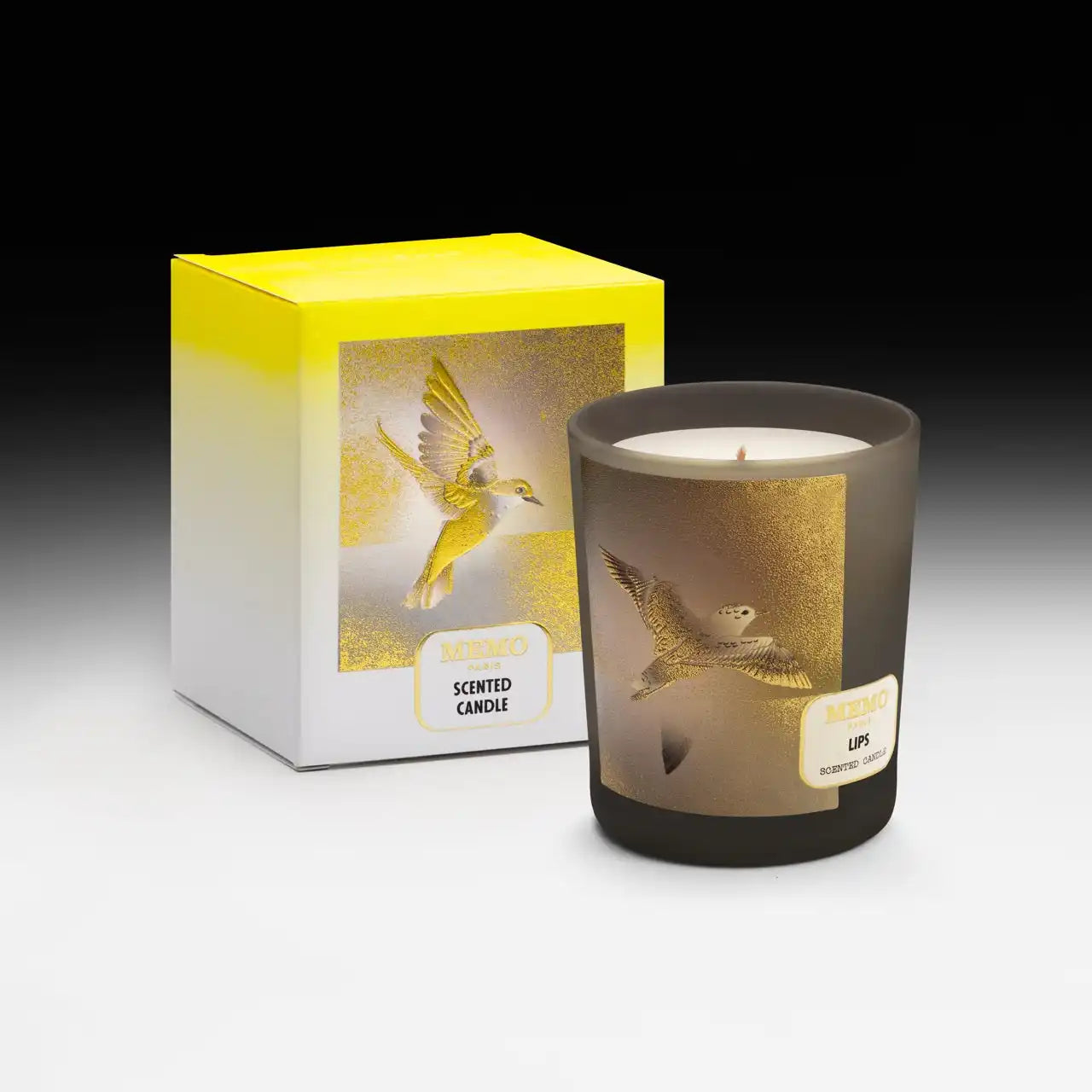 A scented candle in a dark glass jar with a yellow and gold bird design, accompanied by its matching white and yellow box.